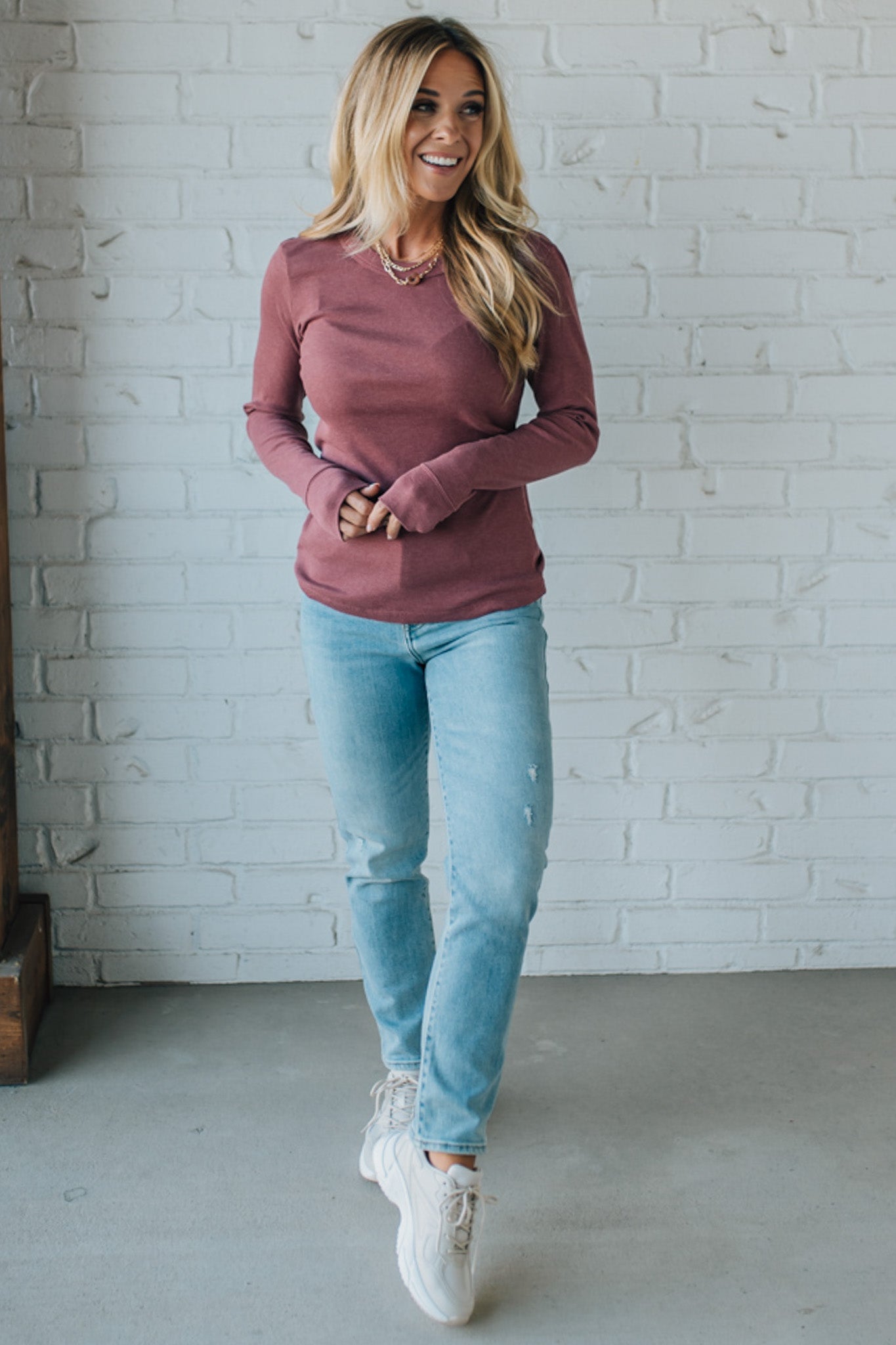Woman wearing a mauve sweatshirt and light blue jeans standing against a white brick wall.