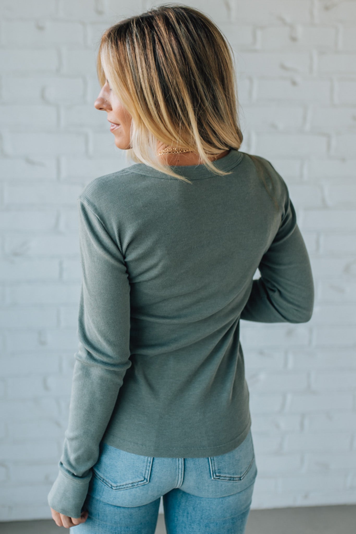 Woman wearing a green long-sleeve top and blue jeans against a white brick wall.