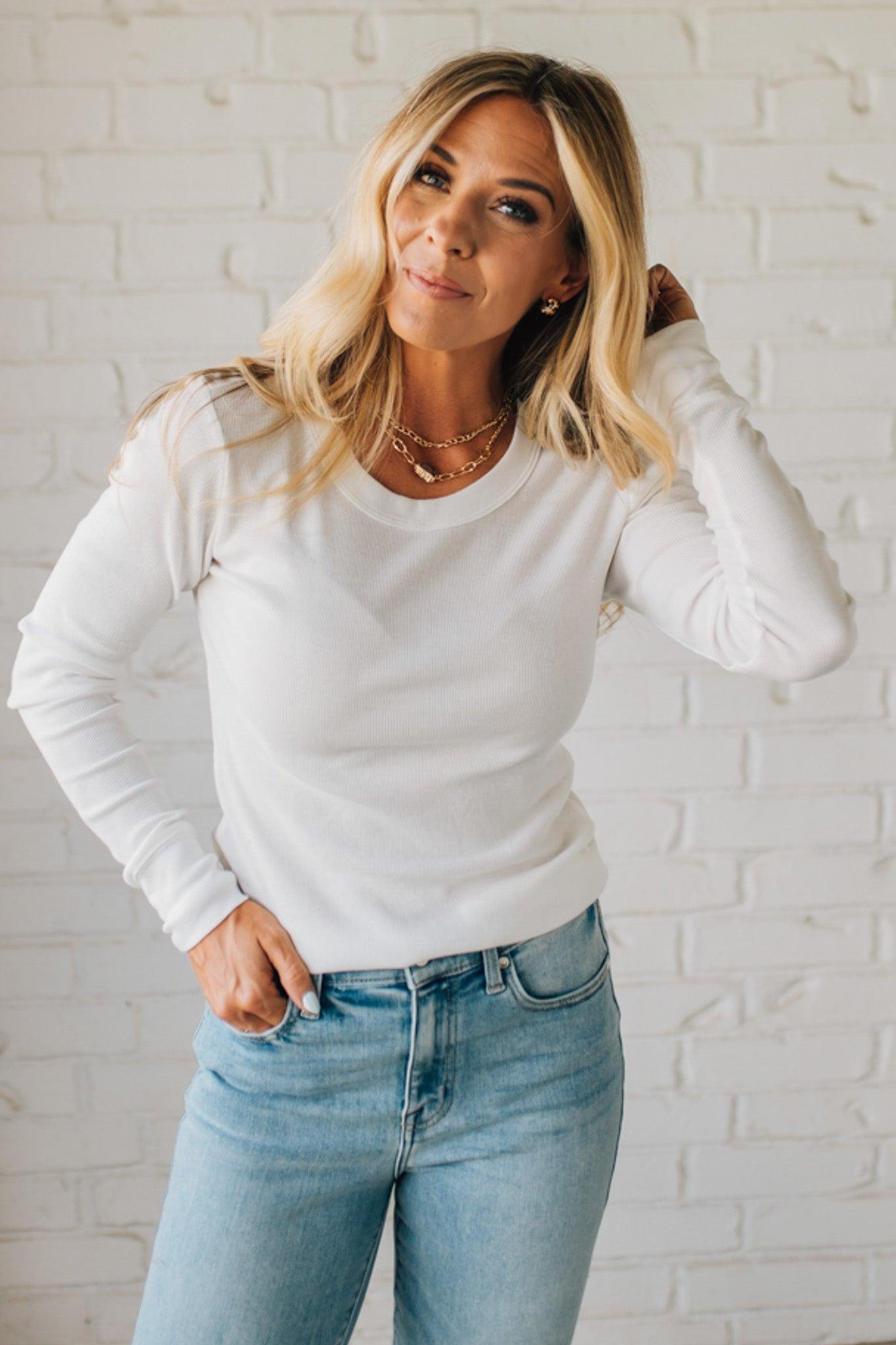 Woman wearing a white sweater and light blue jeans against a white brick wall.