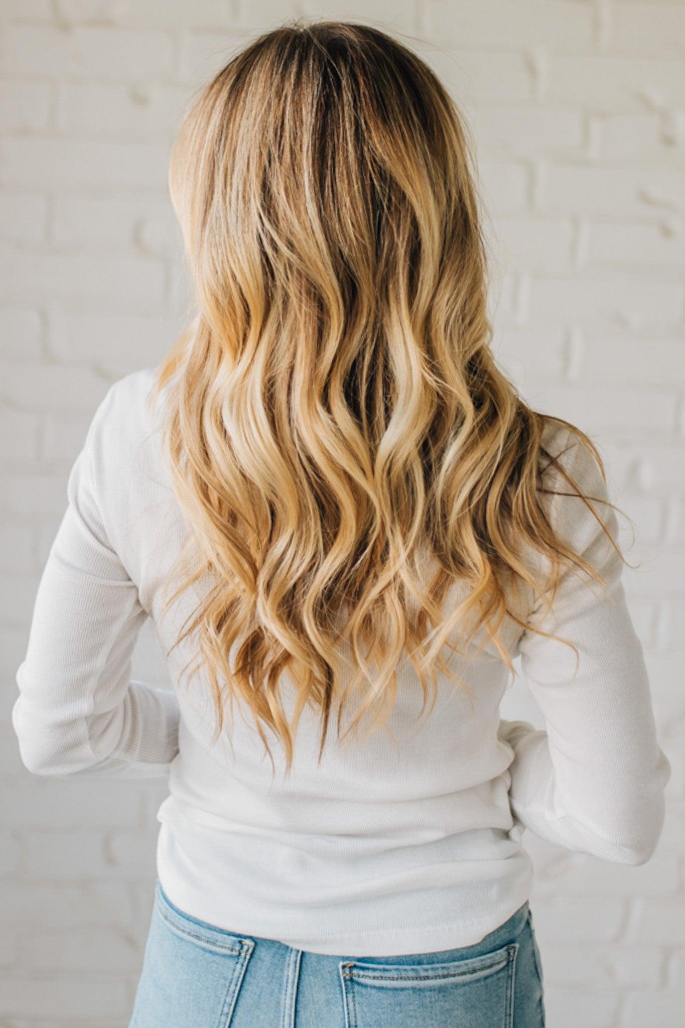 Person with long, wavy blonde hair wearing a white top and blue jeans against a light brick wall.