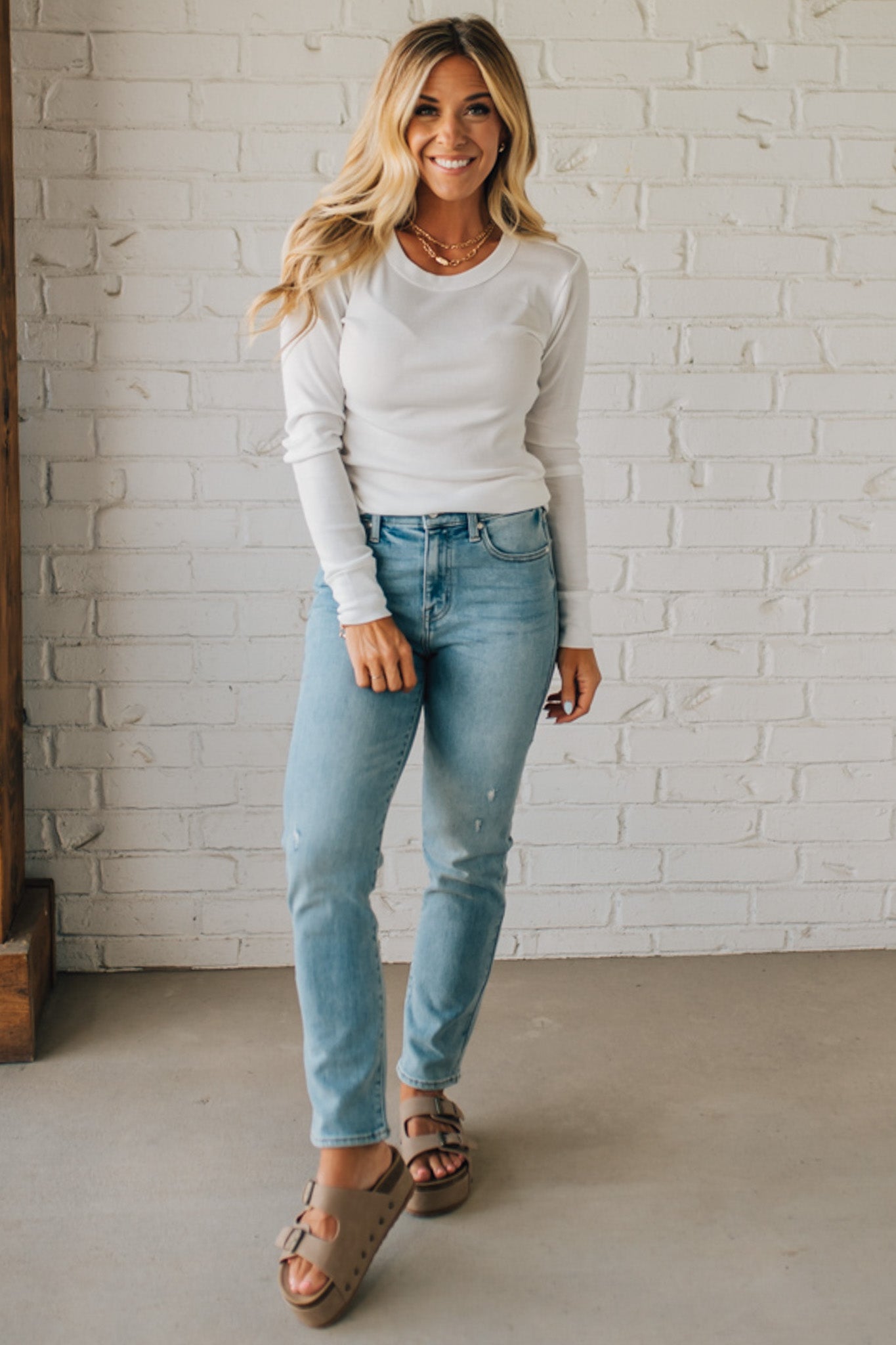 Woman wearing a white sweater and light blue jeans standing against a white brick wall.