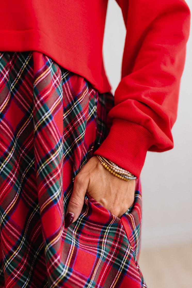 Blonde woman wearing a knee length dress with red french terry top, and red plaid skirt bottom with pockets on the sides.