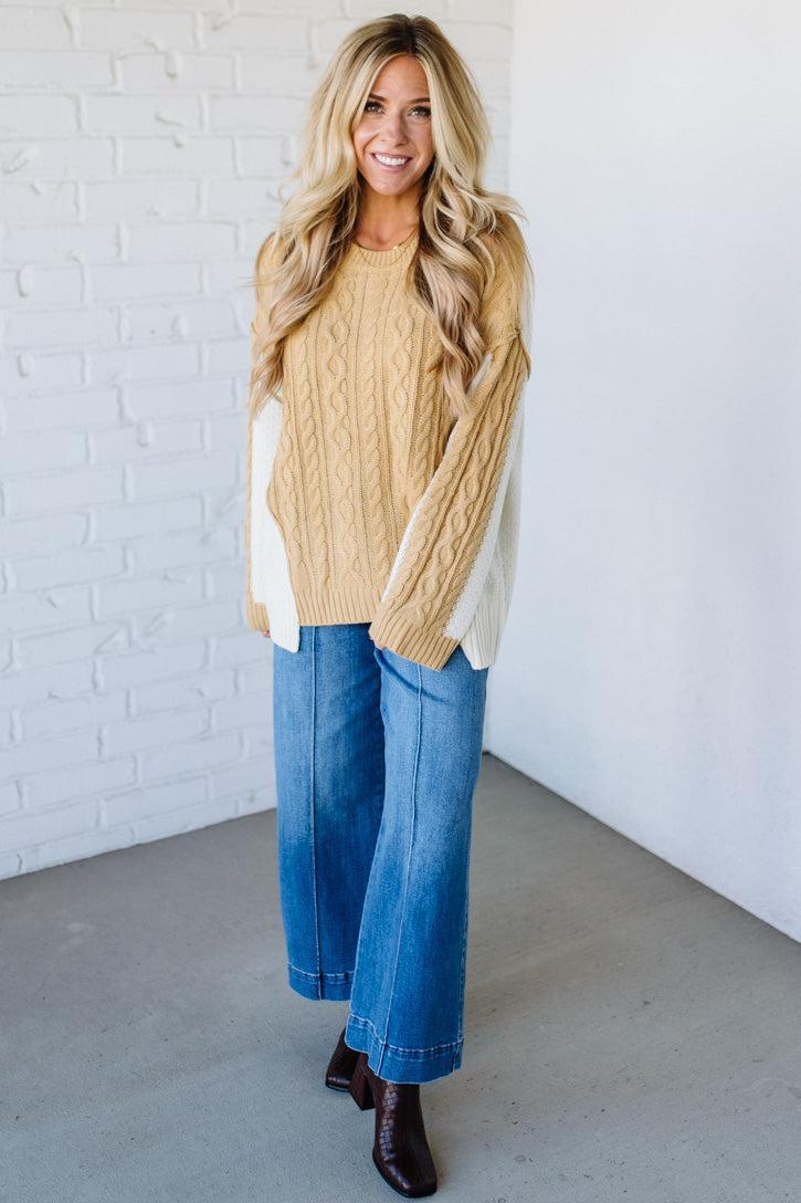 Woman wearing a beige sweater with white accents, blue jeans, and brown shoes against a white brick wall.