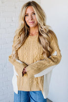 Woman wearing a beige cable knit sweater with white accents against a white brick wall.