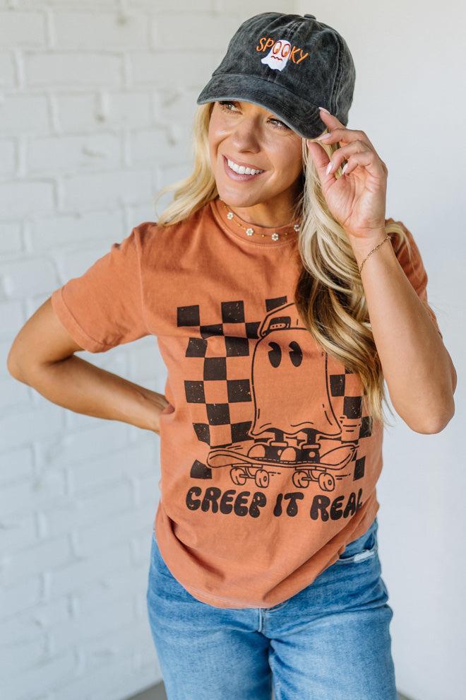 Woman wearing a graphic t-shirt and cap with text, standing against a white brick wall.