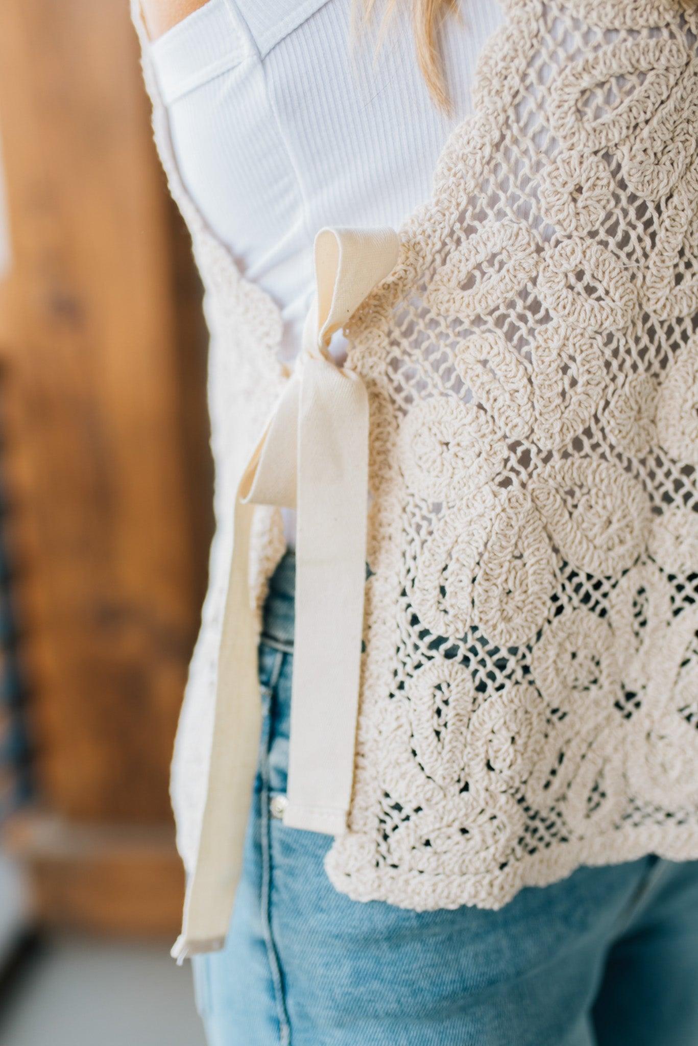 BLONDE WOMAN WEARING A FLORAL CROCHET VEST TOP WITH OPEN SIDES WITH TWILL TAPE TIE.