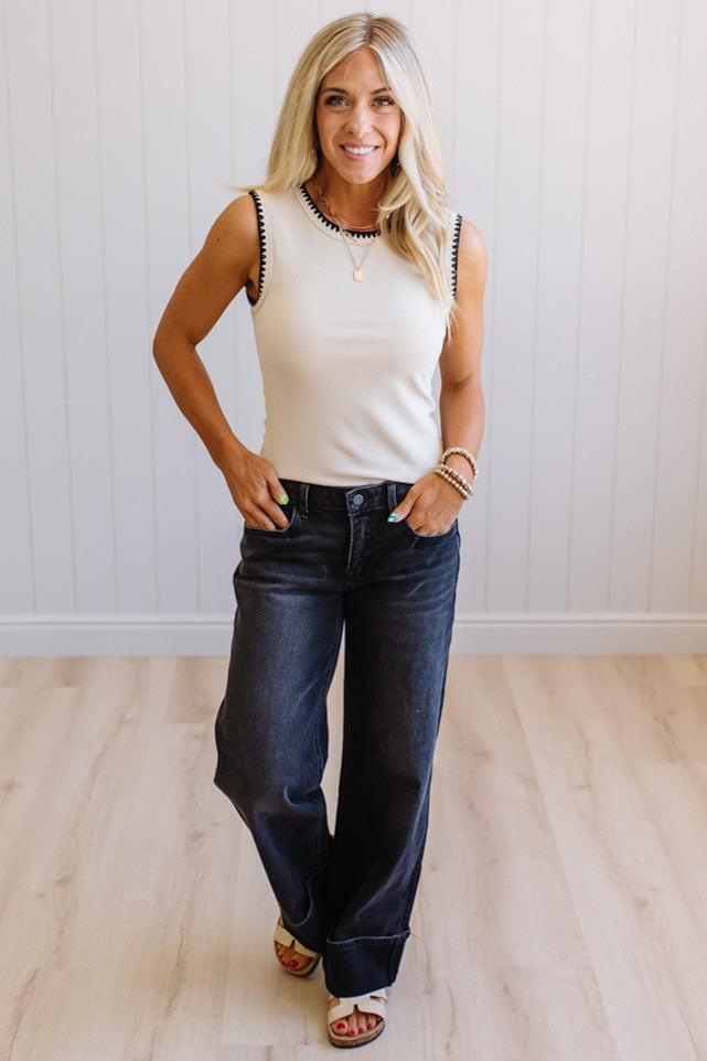 Woman wearing a white sleeveless top and dark blue jeans standing against a light wooden floor and white wall.
