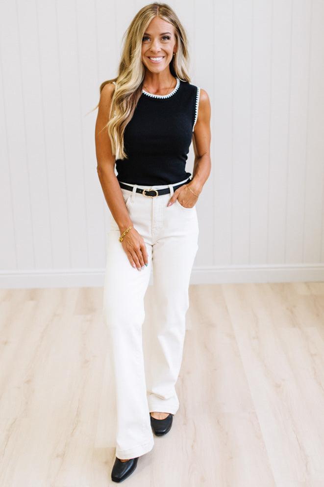 A woman modeling a sleeveless black knit top with white trim, paired with white wide-leg pants and black flats, standing in a bright room with light wood floors and white paneled walls.