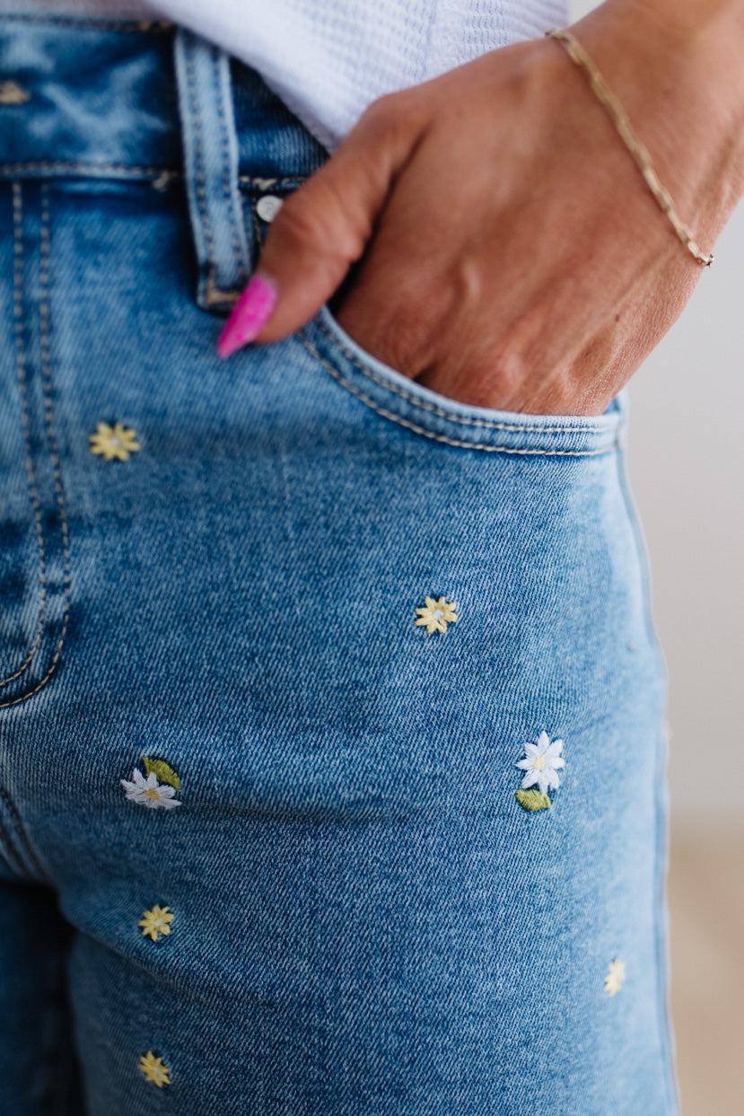 Close-up of blue jeans with floral embroidery and a hand wearing pink nail polish.