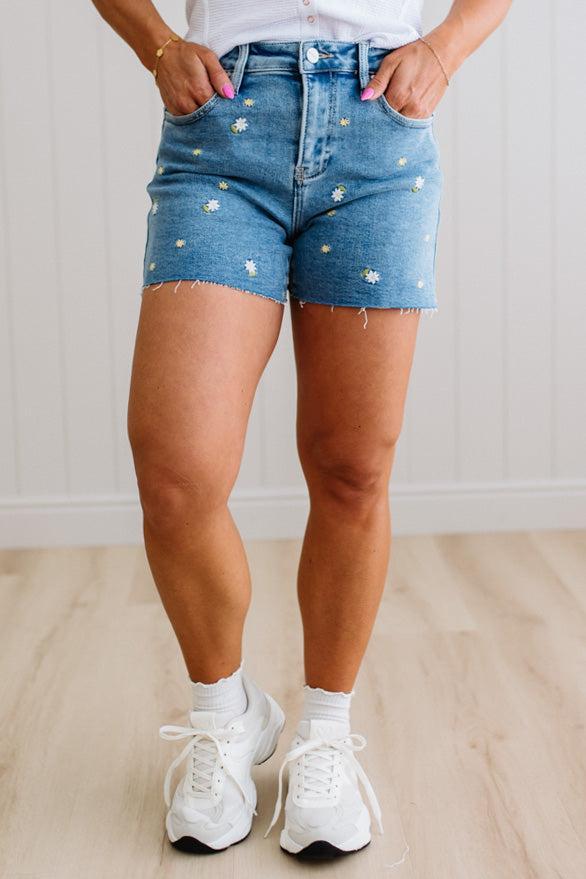 Person wearing blue denim shorts with white embellishments and white sneakers on a light wooden floor.
