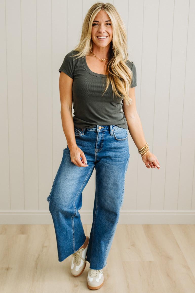 Woman wearing a green t-shirt and blue jeans standing against a light wooden paneled wall.