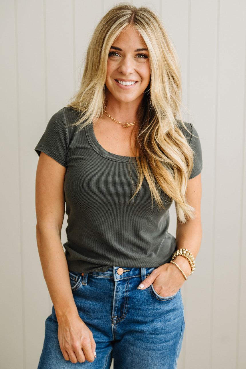 Woman wearing a dark gray t-shirt and blue jeans against a light wooden panel background