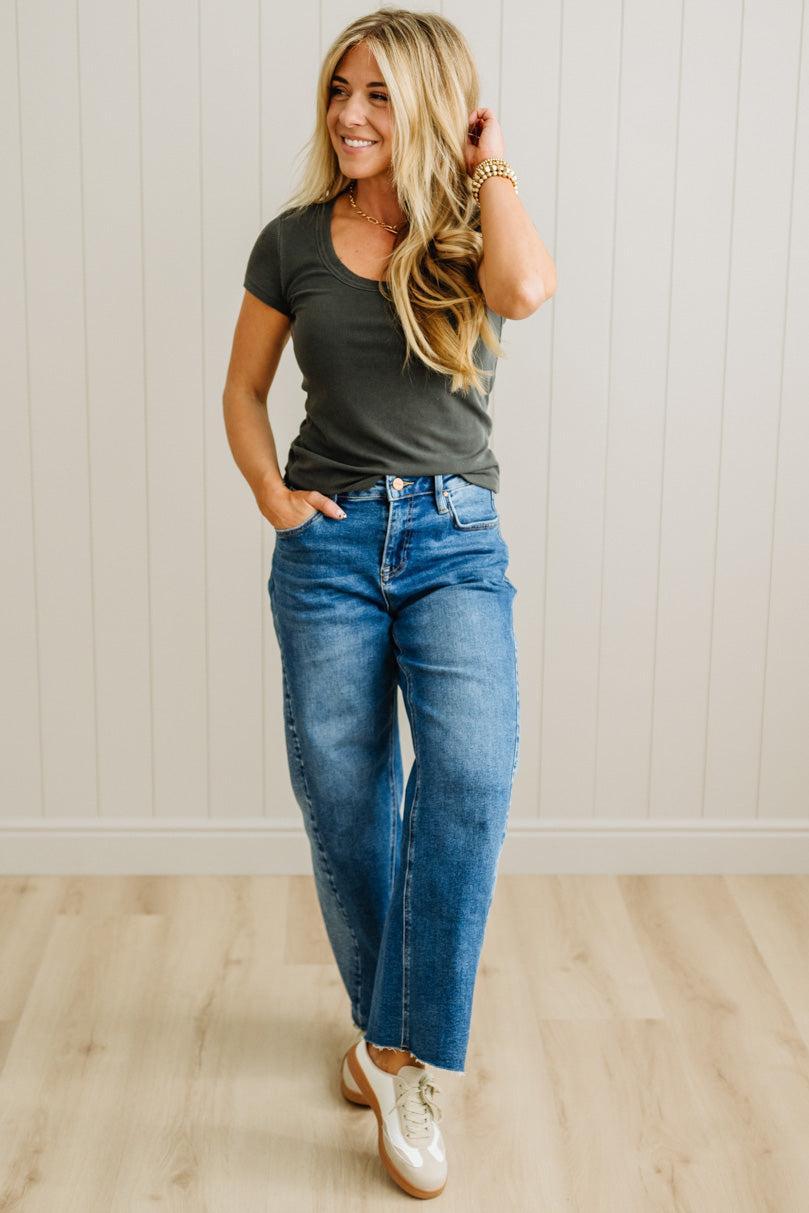 Woman wearing a gray t-shirt and blue jeans standing against a light wooden paneled wall.