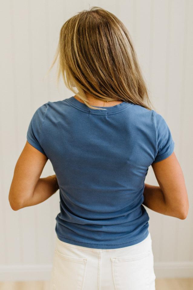 A woman viewed from behind wearing a short-sleeve crew-neck t-shirt in a dusty slate blue color, paired with white pants, set against a light beige paneled wall.