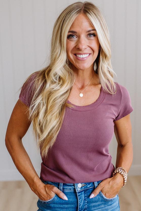 A smiling blonde woman wearing a mauve-purple short-sleeve crewneck t-shirt and blue jeans, standing against a light-colored paneled wall.