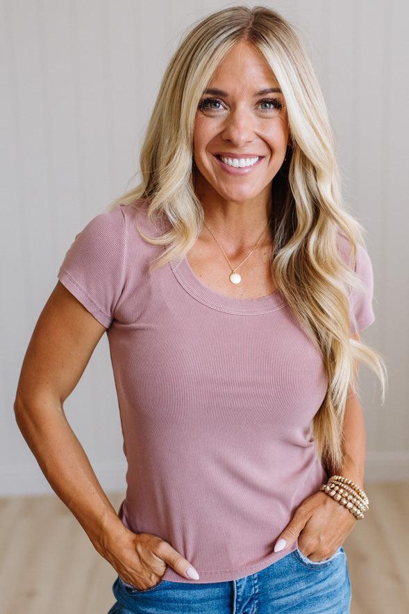 A smiling blonde woman wearing a dusty rose short-sleeve scoop-neck t-shirt and blue jeans, standing with hands on hips against a light neutral background.