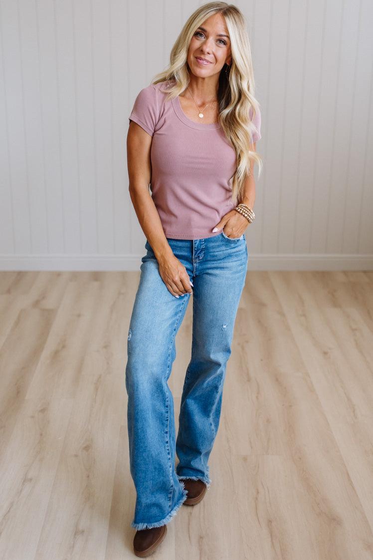 A woman modeling a dusty rose short-sleeve scoop neck t-shirt and light-wash blue flare jeans with raw hems, standing on a light wood floor against a white paneled wall.
