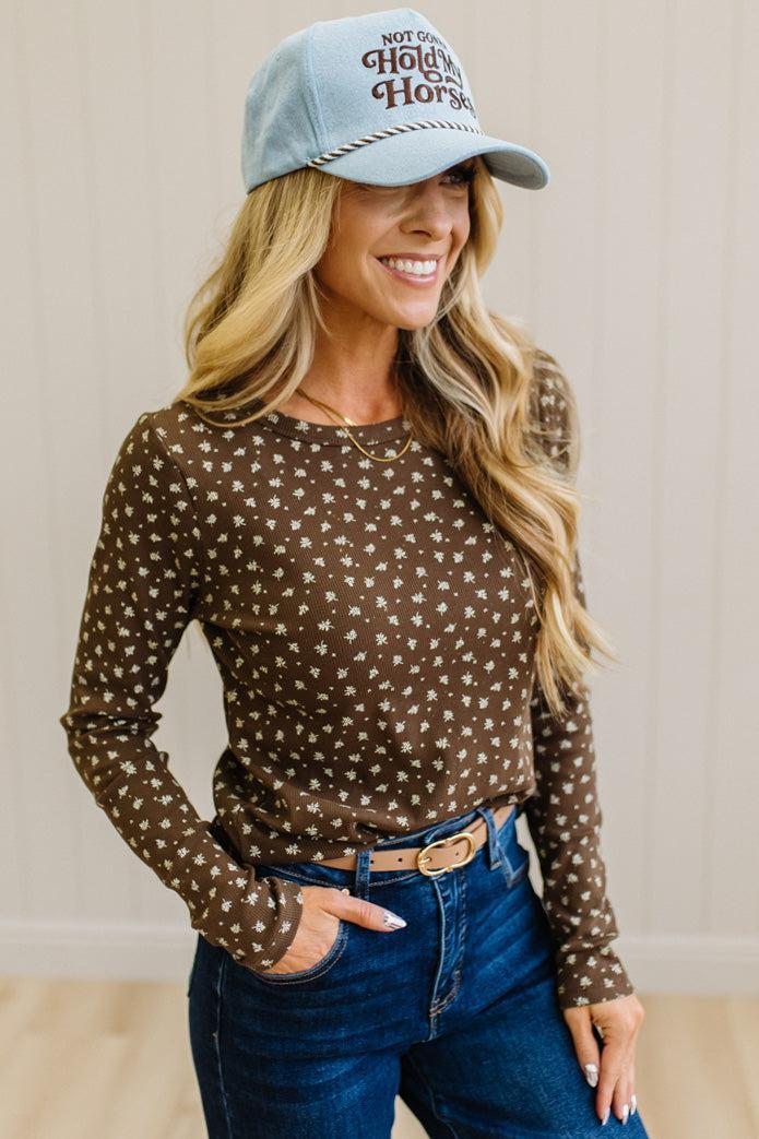 Blonde woman in dark jeans and a light denim ball cap wearing a brown long sleeve tee with botanical print.