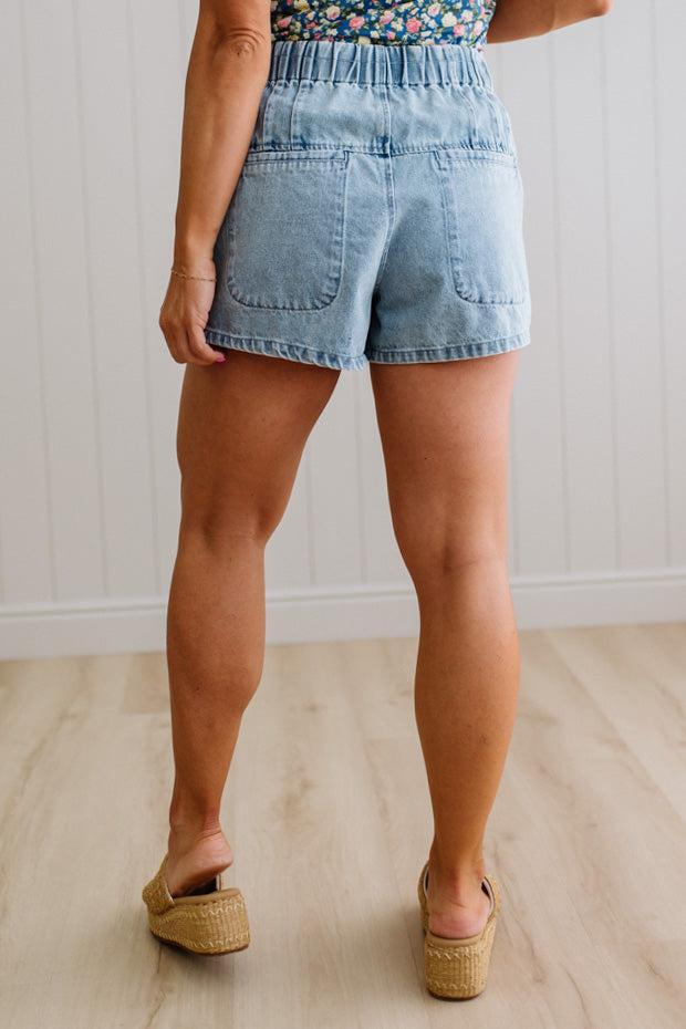Blonde woman in a blue floral tank wearing drawstring waist flare denim shorts, front button up, with pintuck detail, and light washed denim fabric.