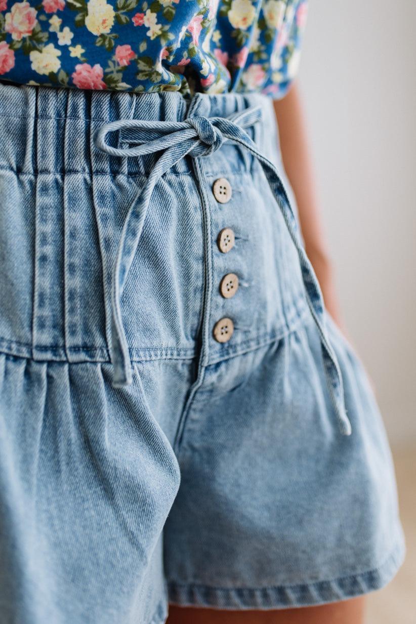 Blonde woman in a blue floral tank wearing drawstring waist flare denim shorts, front button up, with pintuck detail, and light washed denim fabric.