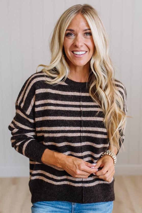 A smiling blonde woman wearing a dark brown and beige horizontally striped knit sweater with blue jeans, standing against a light neutral paneled wall.