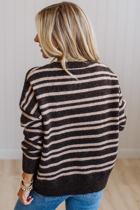 A woman with blonde hair is shown from behind wearing a dark brown and beige horizontally striped knit sweater and blue jeans, standing against a white paneled wall.