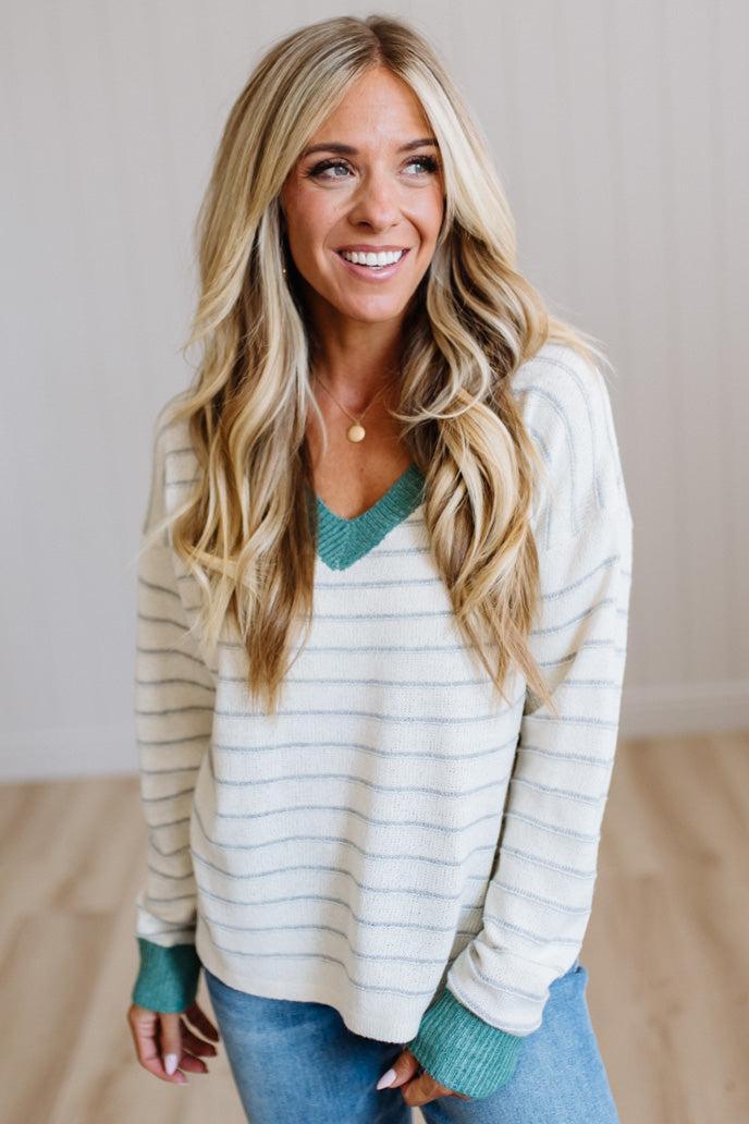 A smiling blonde woman wearing a cream and gray striped knit sweater with teal green trim on the V-neck, cuffs, and hem, paired with blue jeans.