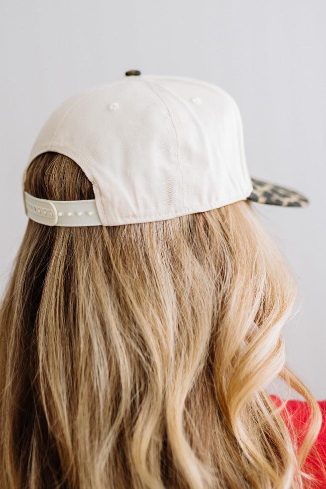 Blonde woman in a red top and gold necklace wearing a cream hat with leopard brim and red embroidered text that says "dear santa just leave your credit card"