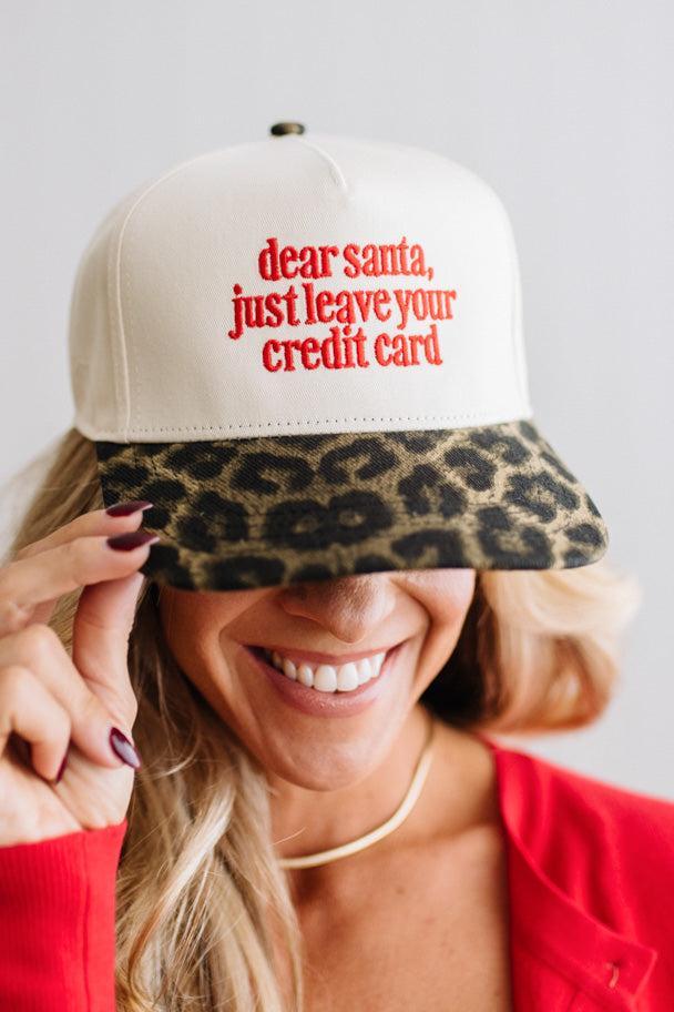 Blonde woman in a red top and gold necklace wearing a cream hat with leopard brim and red embroidered text that says "dear santa just leave your credit card"