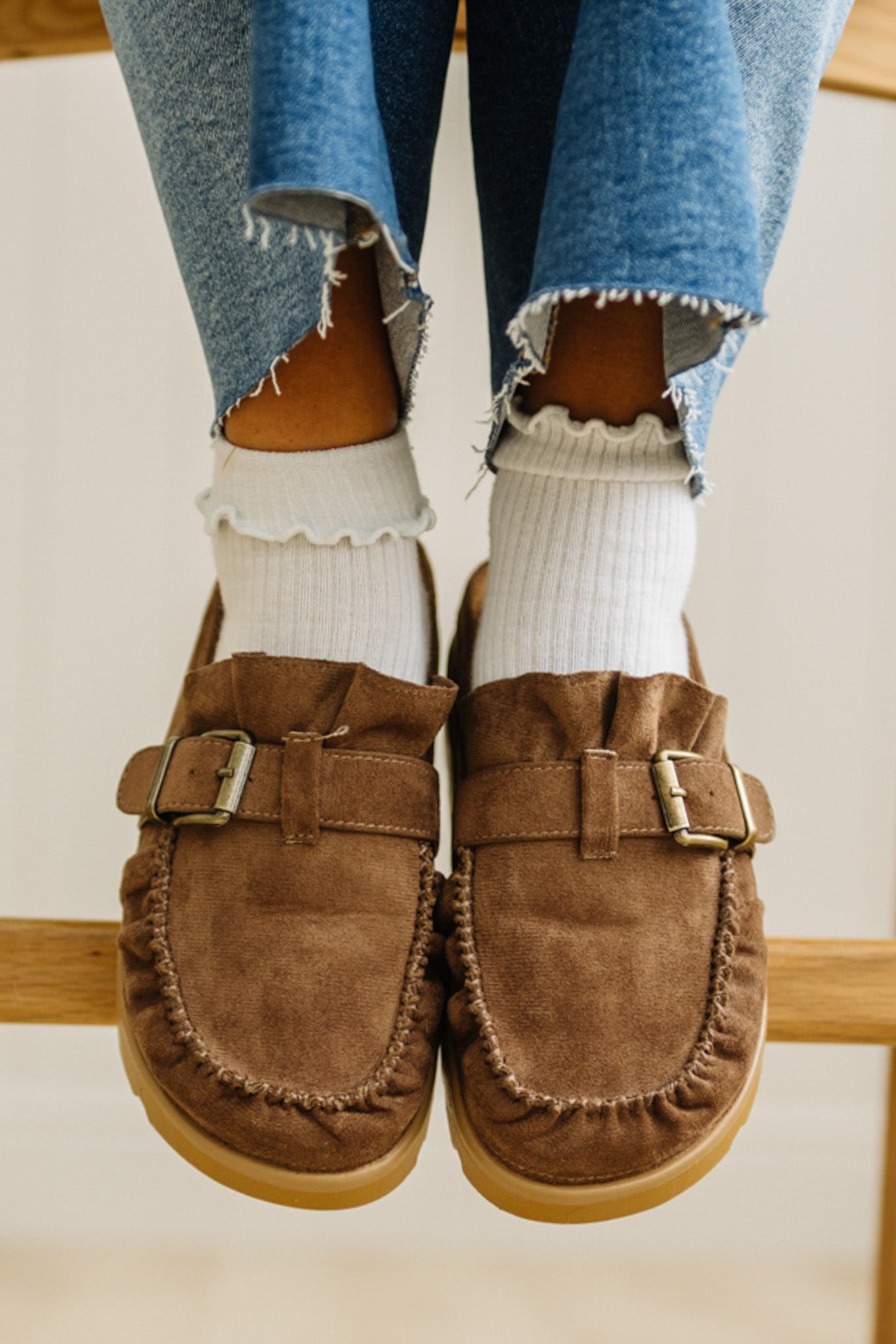 Brown suede loafers worn with white socks and blue jeans on a light background