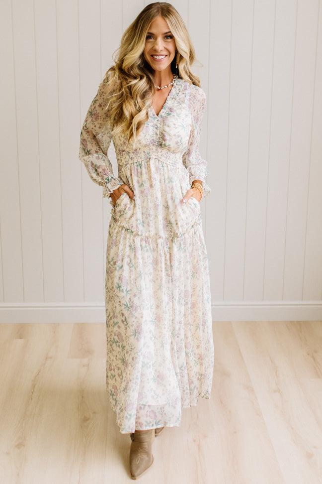 Woman wearing a cream floral dress against a white paneled wall.