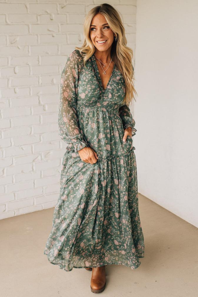 Woman wearing a green floral dress standing against a white brick wall.