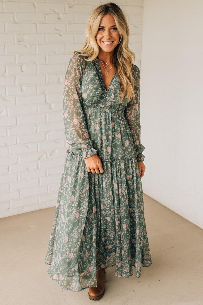 Woman wearing a green floral dress standing against a white brick wall.