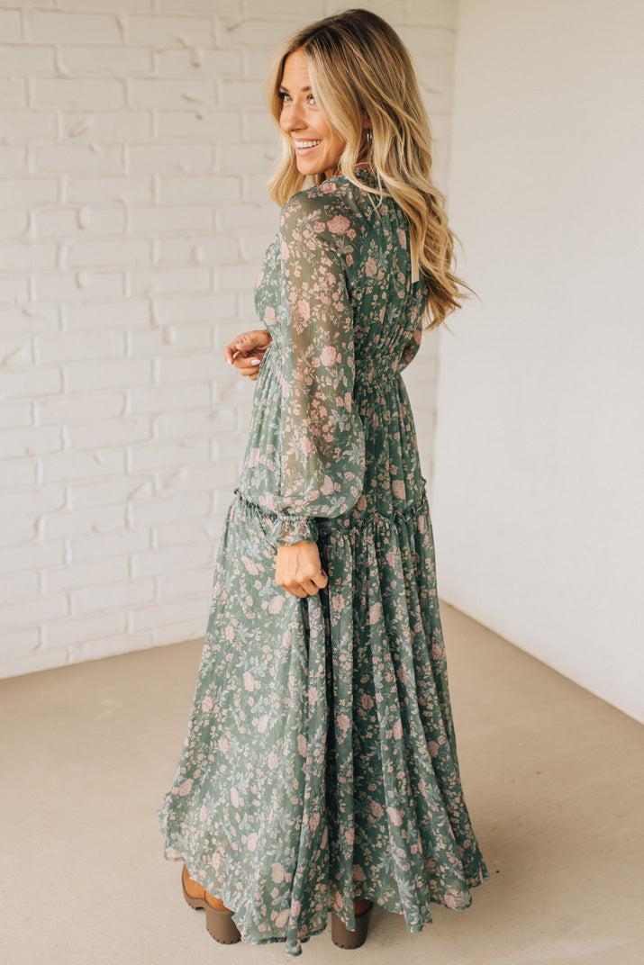 Woman wearing a green floral dress standing against a white brick wall.