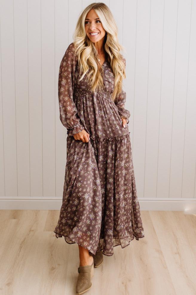 A woman modeling a long-sleeve, tiered maxi dress with a brown and beige floral pattern, standing in front of a white paneled wall and light wood floor.