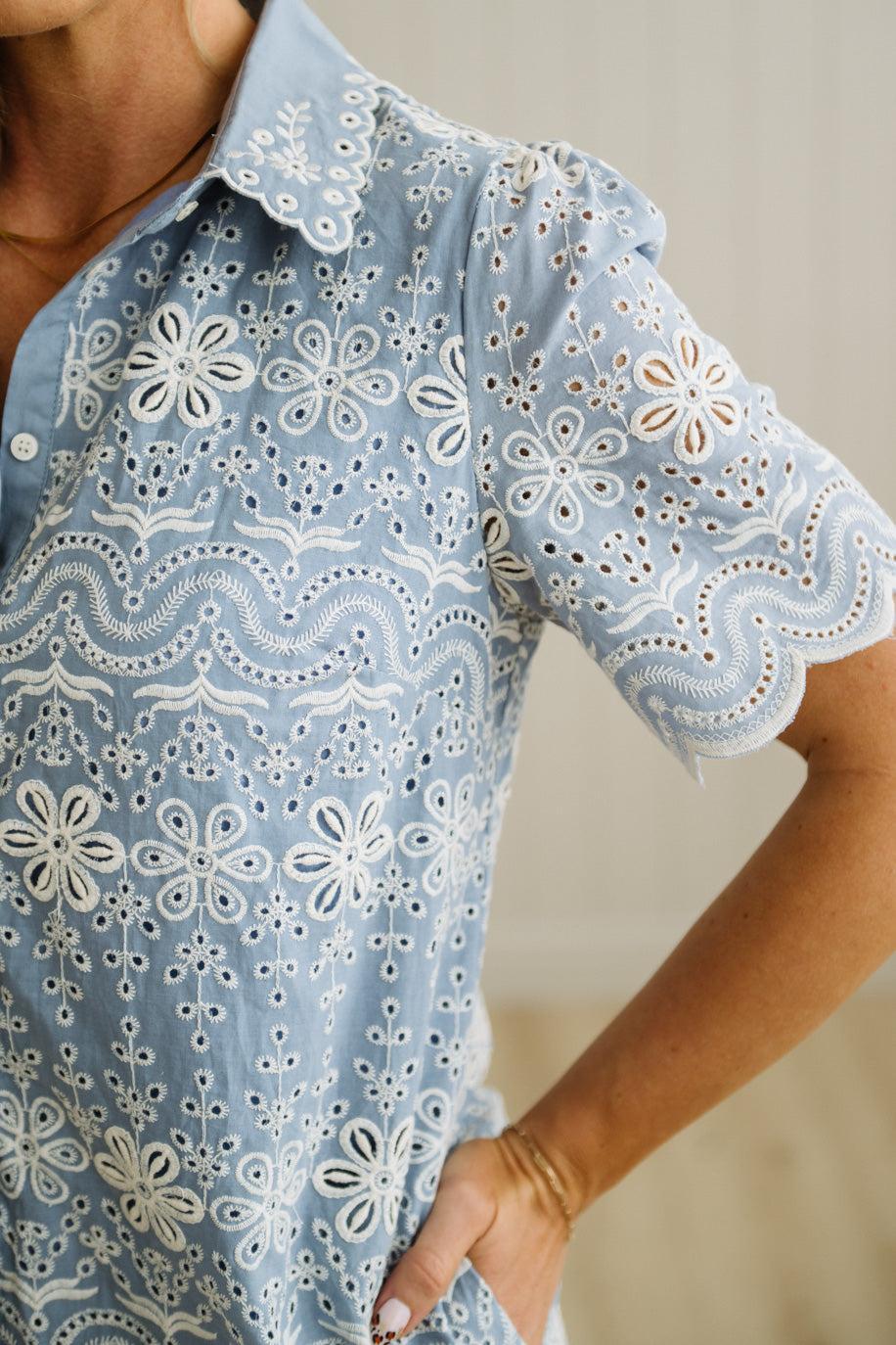 Blonde woman wearing a button front collared shift dress with scalloped edges, and all over eyelet lace embroidery in light blue chambray.