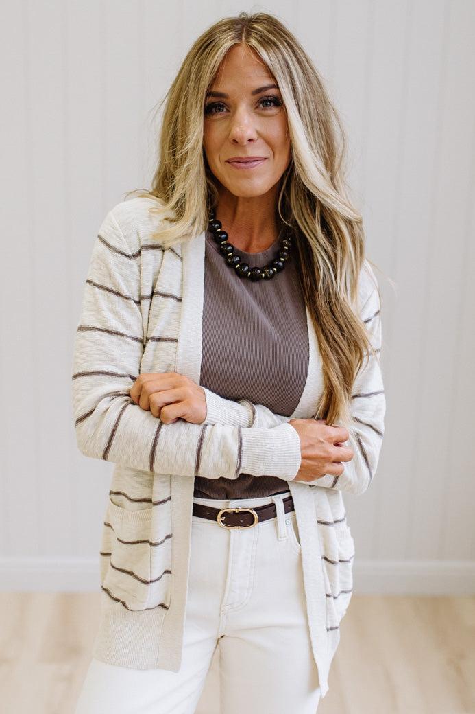 A woman with long blonde hair stands indoors wearing a cream and brown striped cardigan over a taupe top, white pants, and a black beaded necklace.