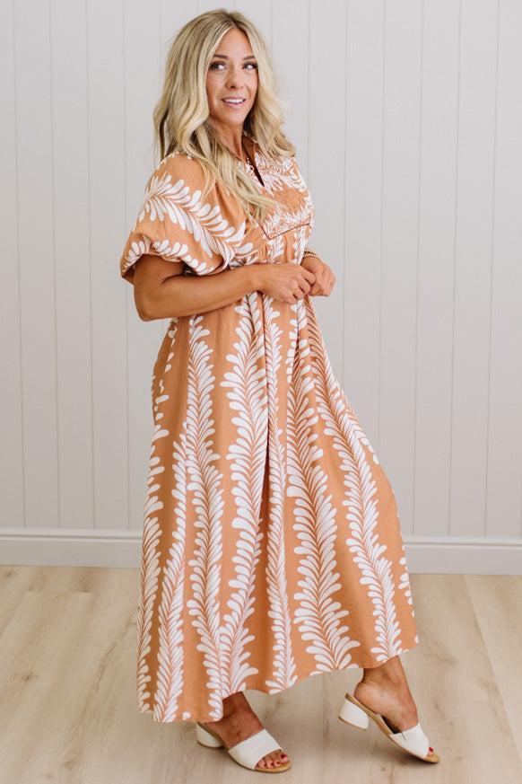 Woman wearing a long, patterned dress standing against a white wall.