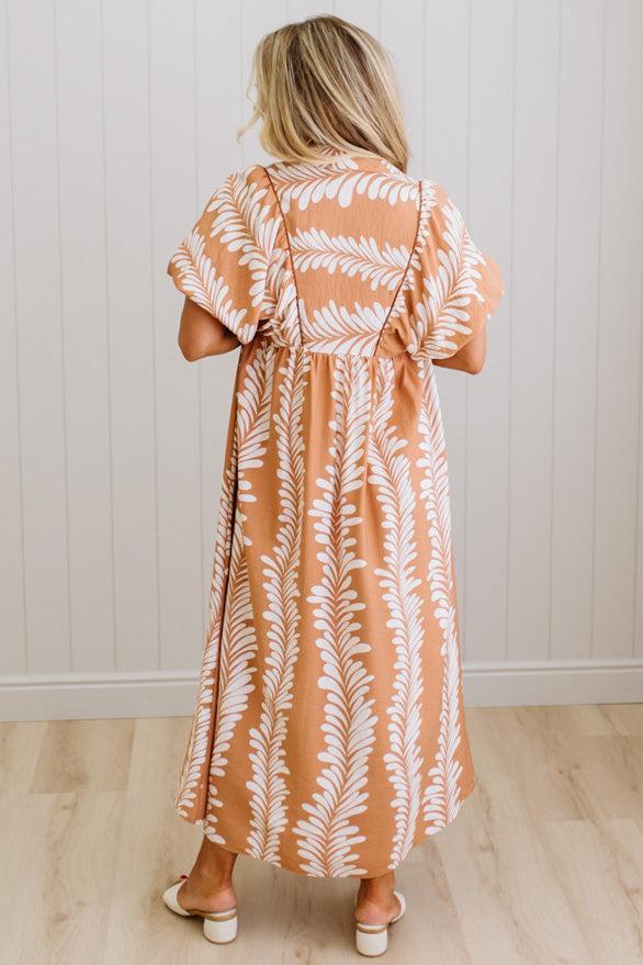 Woman wearing a long dress with a leaf pattern in a room with a white wall and wooden floor.