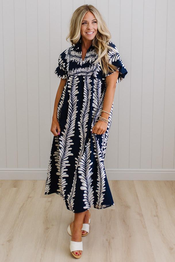 Woman wearing a navy and white patterned dress against a light wooden floor and white paneled wall.
