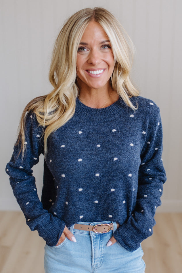 A smiling blonde woman wearing a navy blue knit sweater with small white polka dots, paired with light blue jeans and a brown belt, standing against a light beige wall.