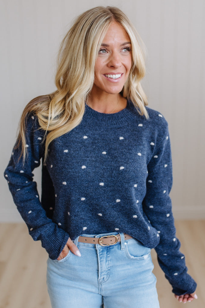 A smiling blonde woman wearing a navy blue knit sweater with small white polka dots, paired with light blue jeans and a brown belt, standing against a light neutral wall.