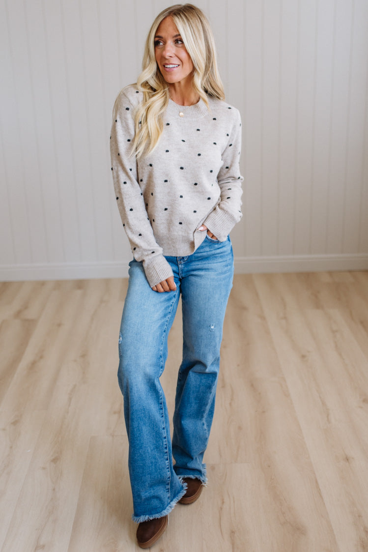 A woman stands in a bright room with light wood flooring and paneled walls, wearing a cream knit sweater with small black polka dots and blue jeans.