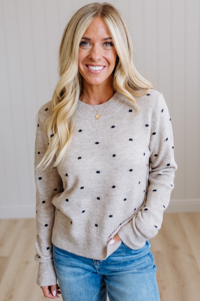 A smiling blonde woman wearing a light beige crewneck sweater with small black polka dots and blue jeans, standing in a brightly lit room with light wood floors and white paneled walls.