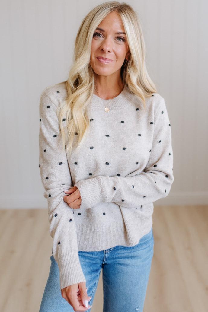 A woman with blonde hair wearing a light beige knit sweater with small black polka dots and blue jeans, standing in a brightly lit room with light wood floors.