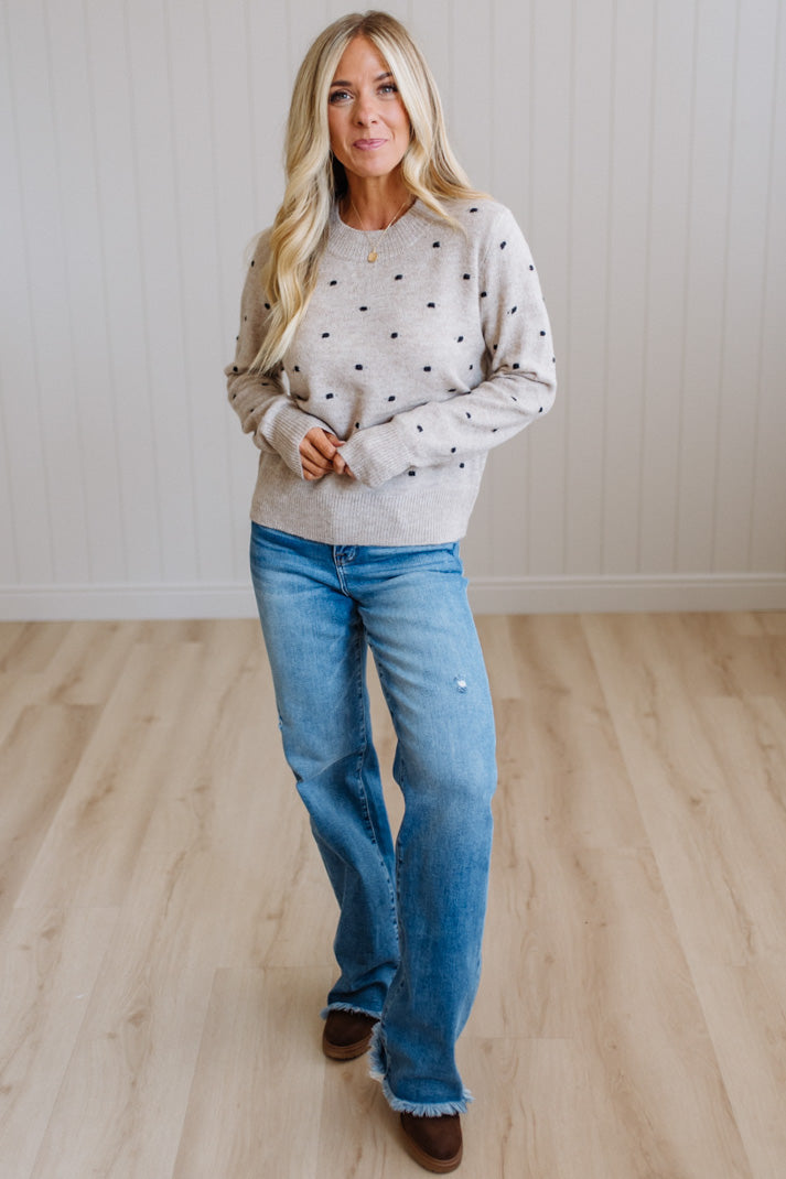 A woman modeling a light beige crewneck sweater with small black polka dots, paired with blue jeans and brown boots, standing in a room with light wood floors and white paneled walls.