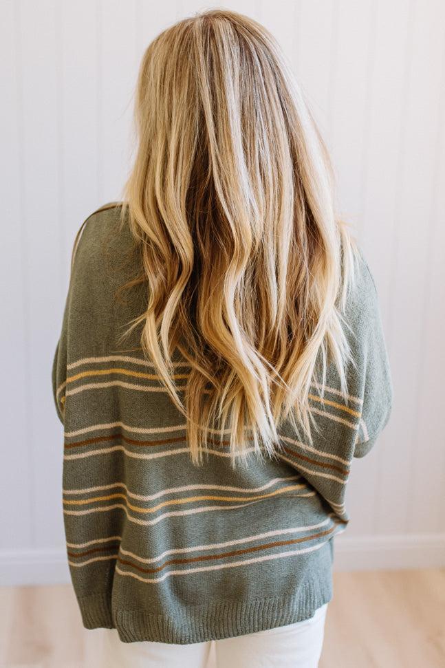 A woman viewed from behind wearing a loose olive green knit sweater with thin horizontal tan and white stripes, paired with white pants, set against a light-colored paneled wall.