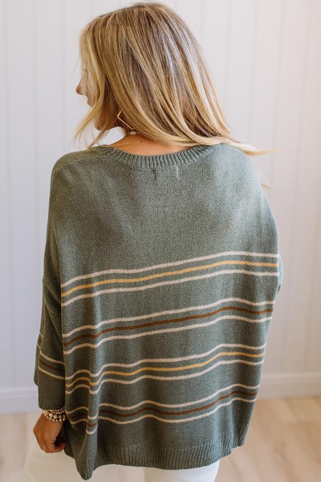 A woman viewed from behind wearing a loose knit sweater in muted sage green with horizontal stripes in tan, cream, and brown, set against a white paneled wall.