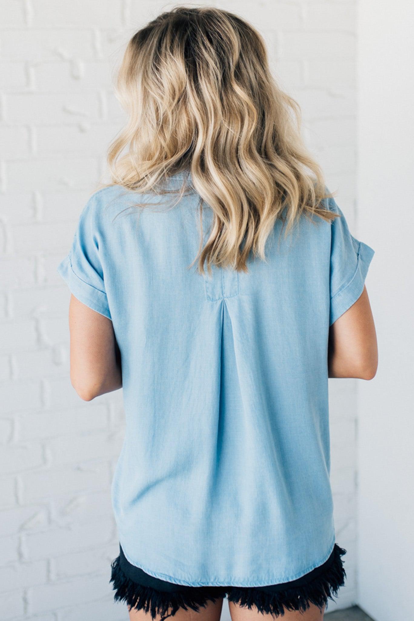 A woman smiling and wearing a blue chambray button front top with a patch chest pocket, dolman shoulders, and short sleeves rolled up to the elbows.