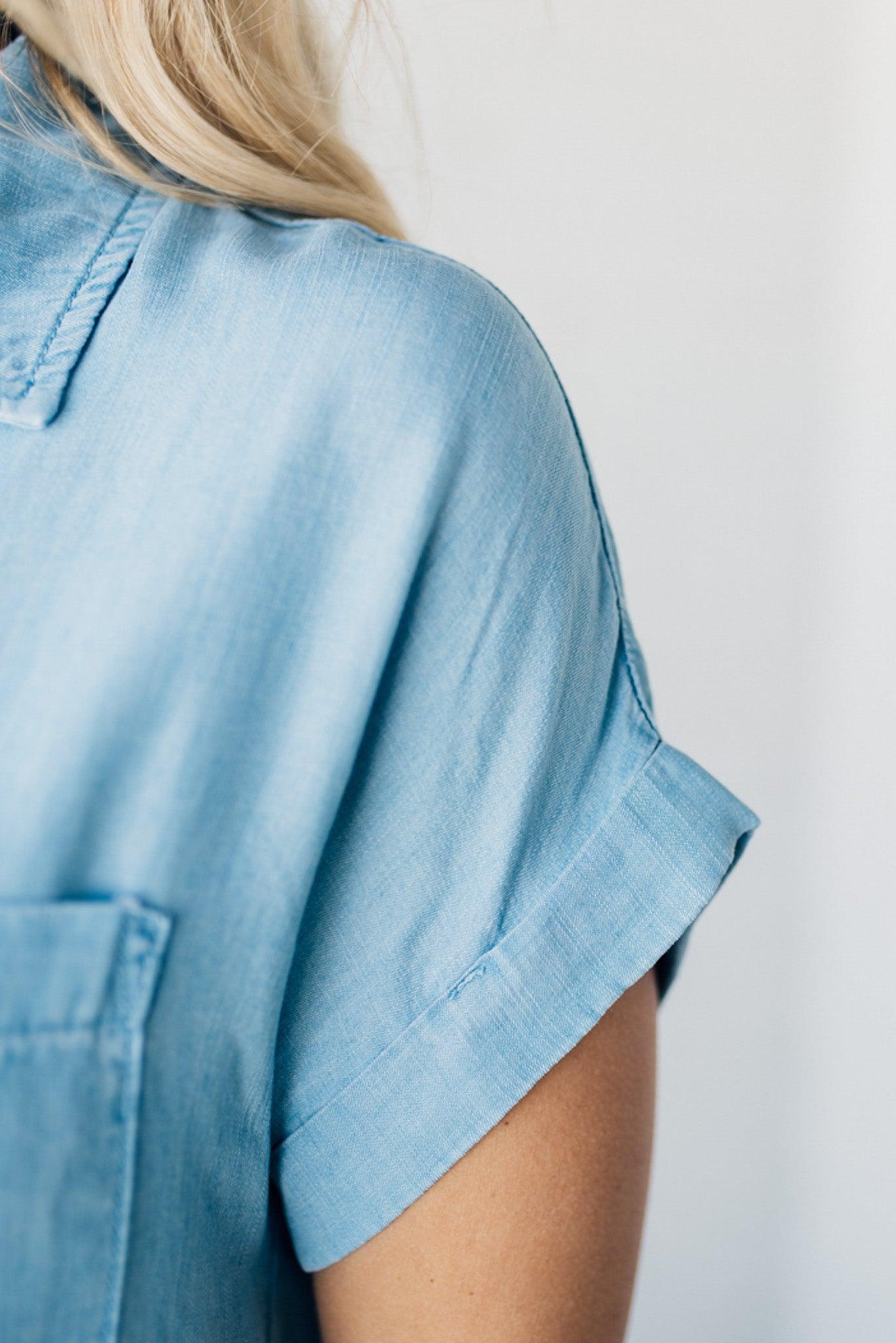 A woman smiling and wearing a blue chambray button front top with a patch chest pocket, dolman shoulders, and short sleeves rolled up to the elbows.